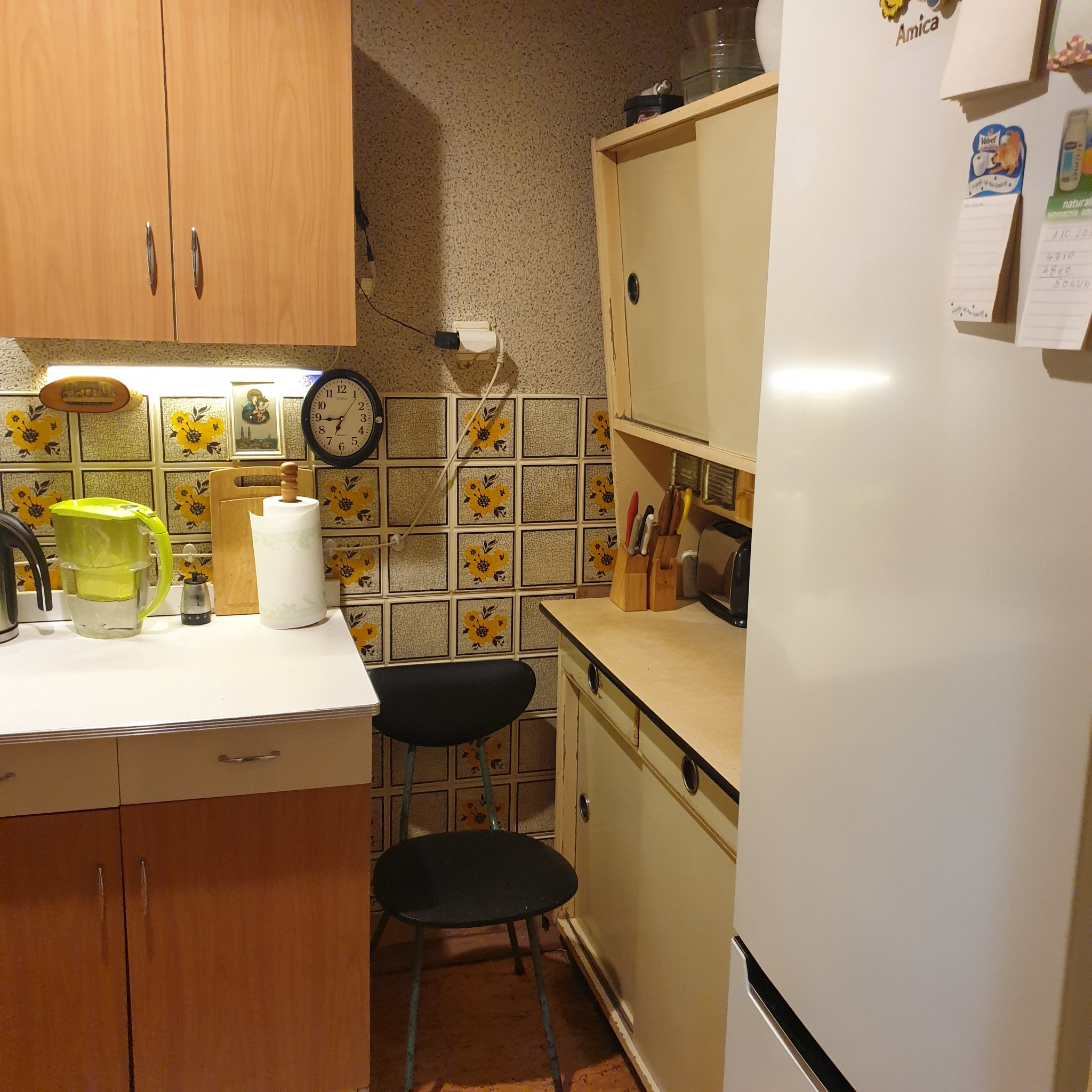 Kuchnia z lat 80-tych z żółtymi kwiatowymi kafelkami, drewnianymi szafkami i białym blatem, na którym stoi filtr do wody, obok stoi czarny stołek i biała lodówka z magnesami.