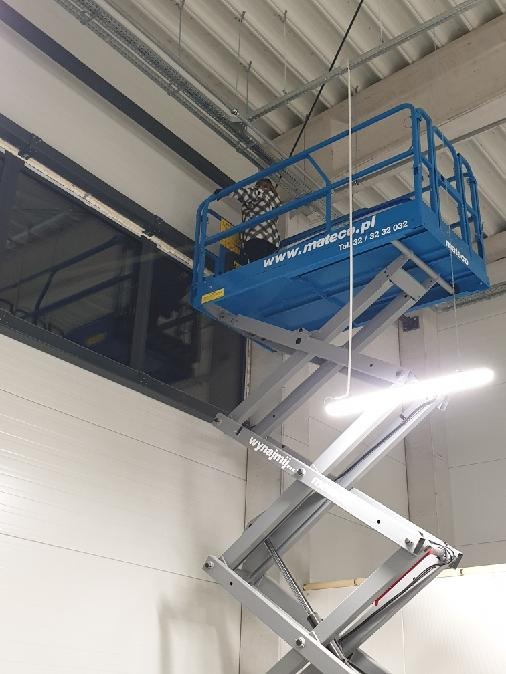 Pracownik w koszu podnośnika nożycowego Mateco, na wysokości, przy oknie w hali przemysłowej z widocznymi instalacjami pod sufitem.