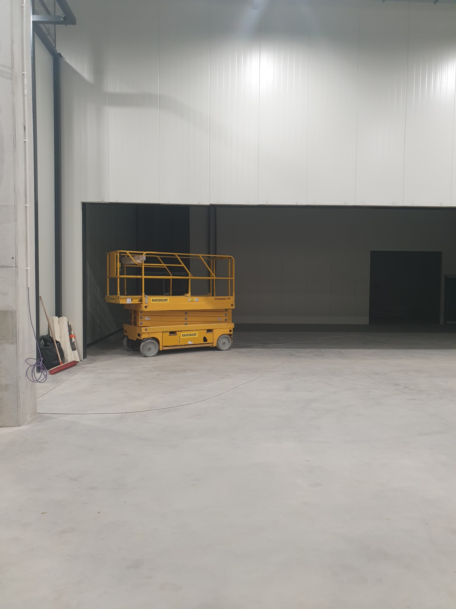 Żółty podnośnik nożycowy Ramirent w pustej hali przemysłowej z betonową podłogą i białymi ścianami z blachy trapezowej; w tle otwarty wjazd do hali.
