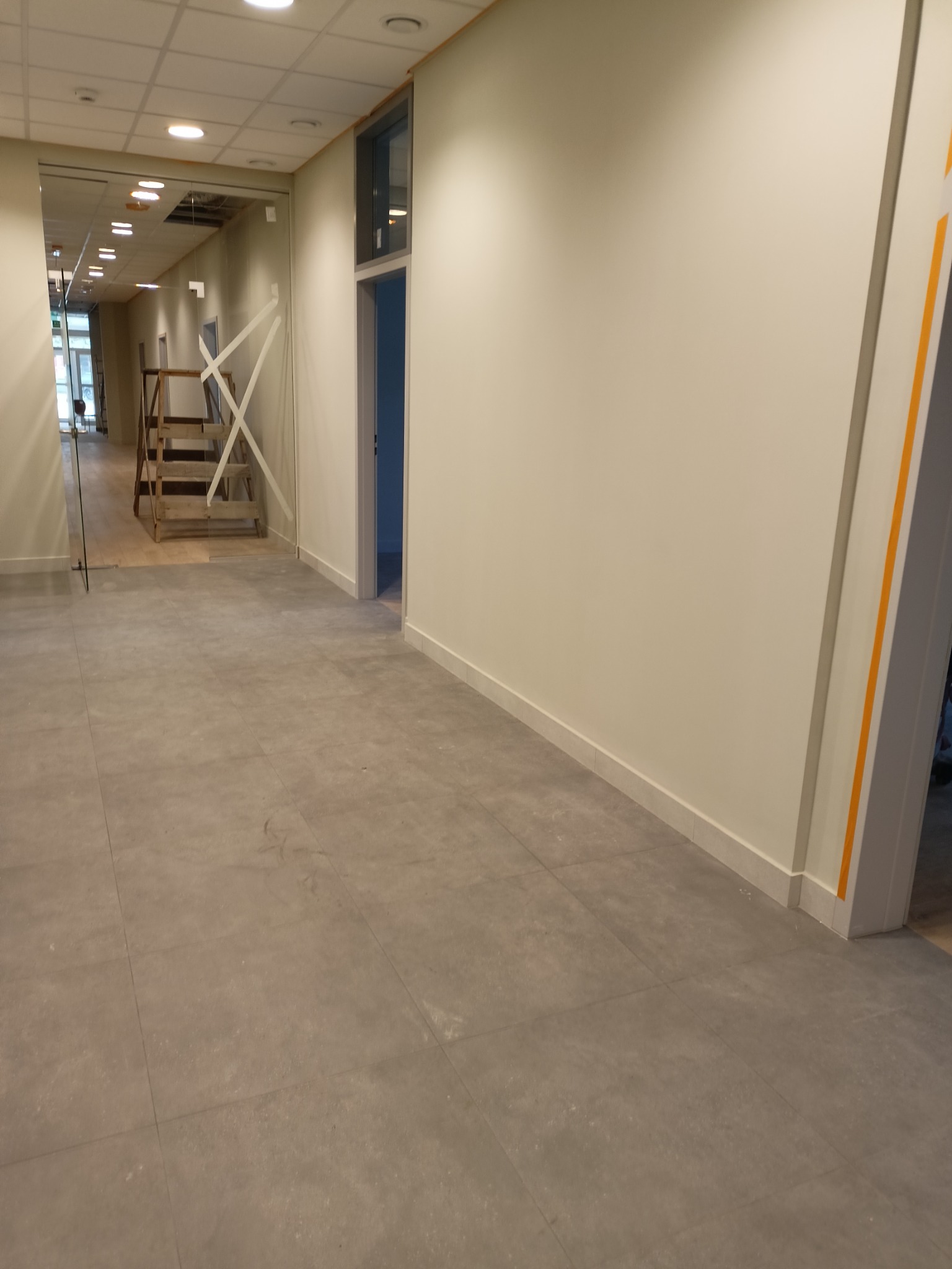 Wnętrze lokalu po remoncie z widocznymi śladami prac budowlanych: szare płytki podłogowe z pozostałościami po budowie, ściany pomalowane na jasny kolor, widoczne rusztowania w głębi pomieszczenia.