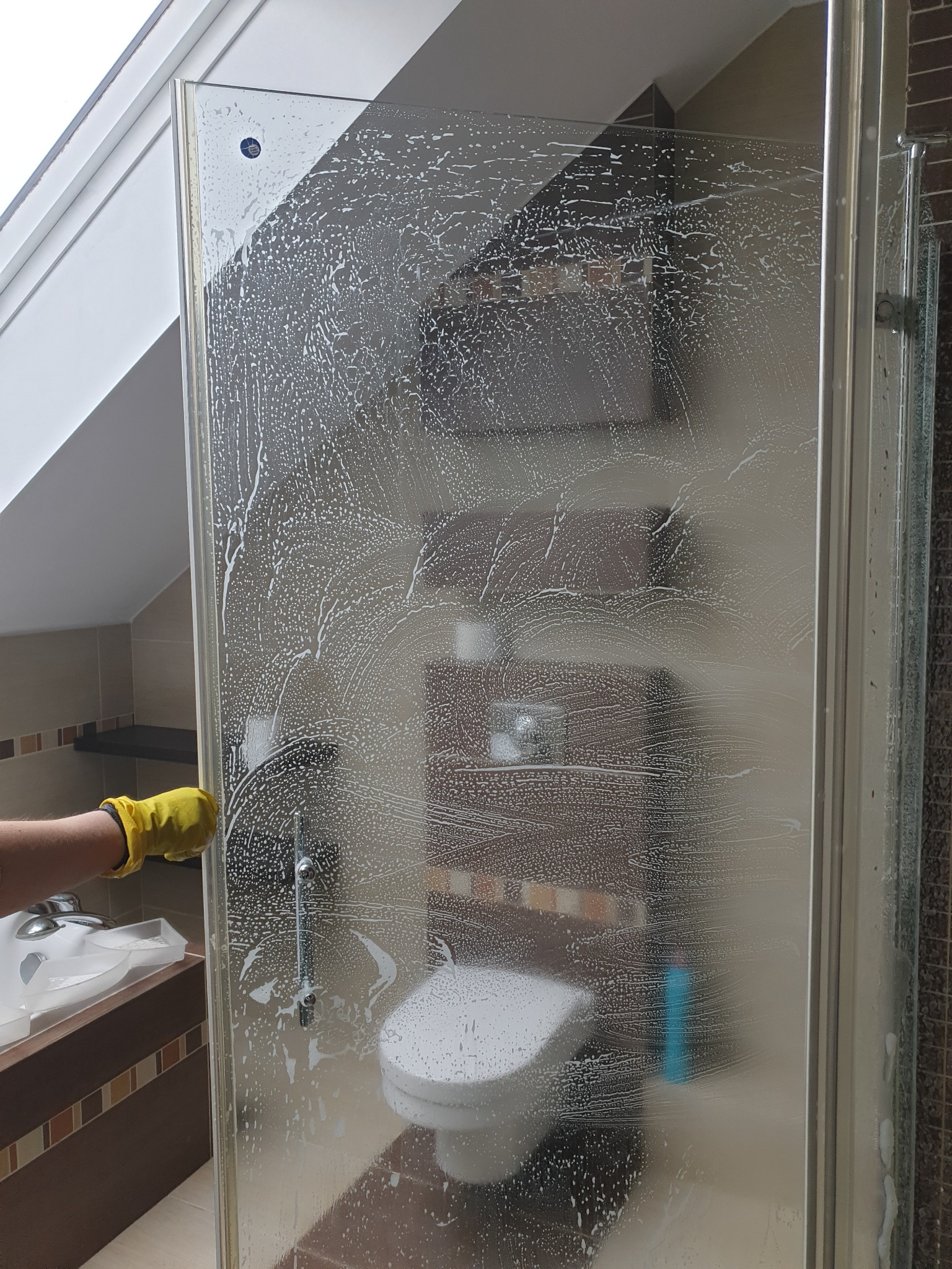 Osoba w żółtej rękawicy czyści kabinę prysznicową za pomocą myjki, pozostawiając ślady piany na szkle. W tle widoczna toaleta i umywalka.