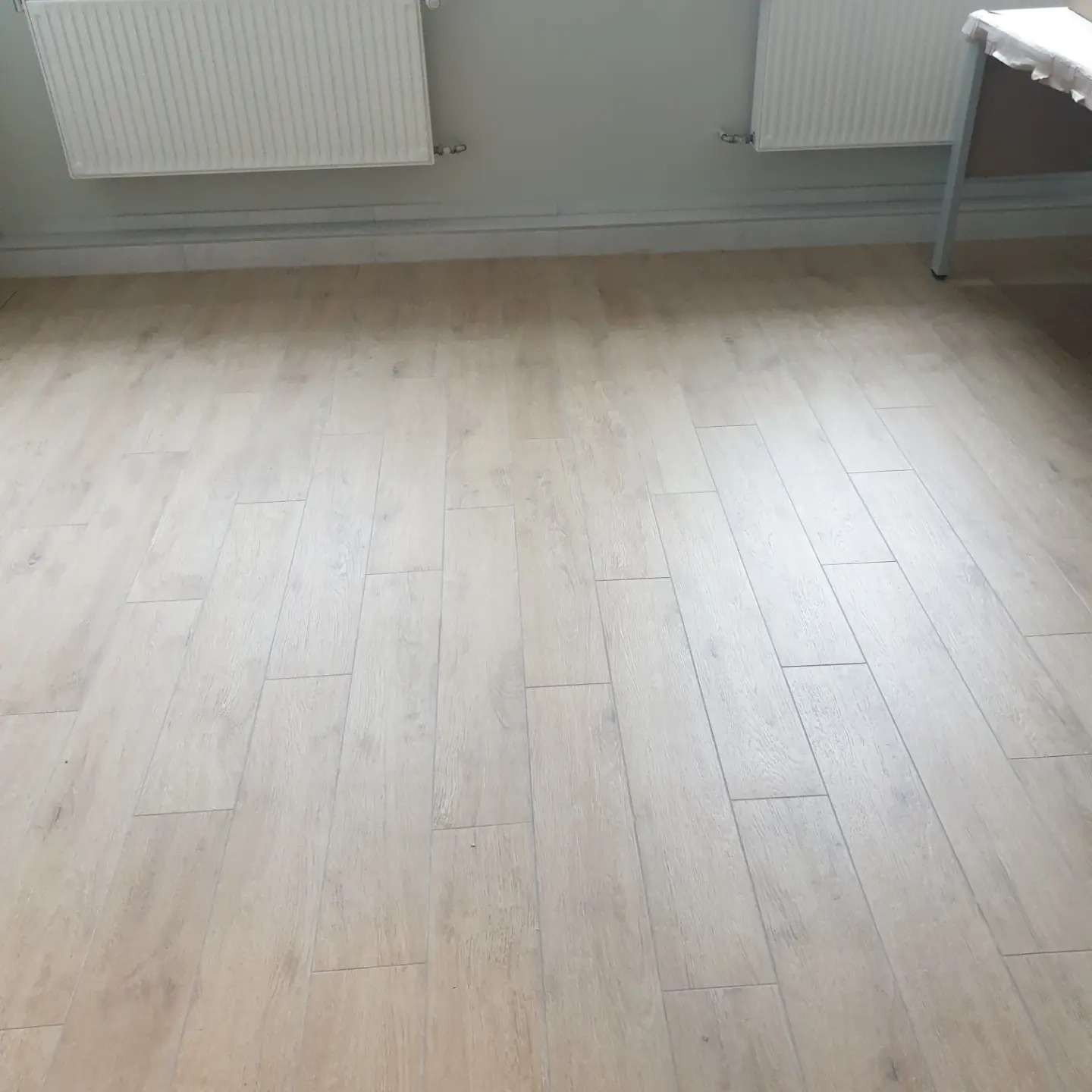 Jasna, drewnopodobna podłoga z paneli ułożonych wzdłuż, dwa białe grzejniki na ścianie i fragment białego stołu w prawym górnym rogu.