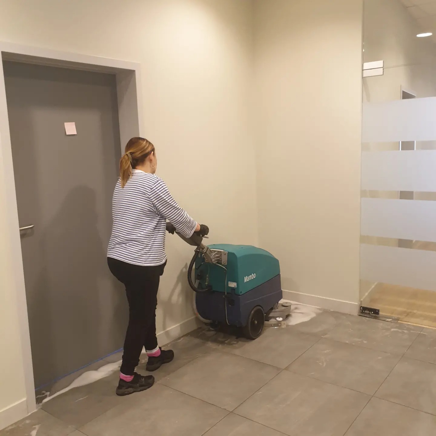 Kobieta w bluzie w paski czyści podłogę w biurze za pomocą profesjonalnej maszyny czyszczącej Mambo, widoczne ślady piany i wilgoci na szarych płytkach.