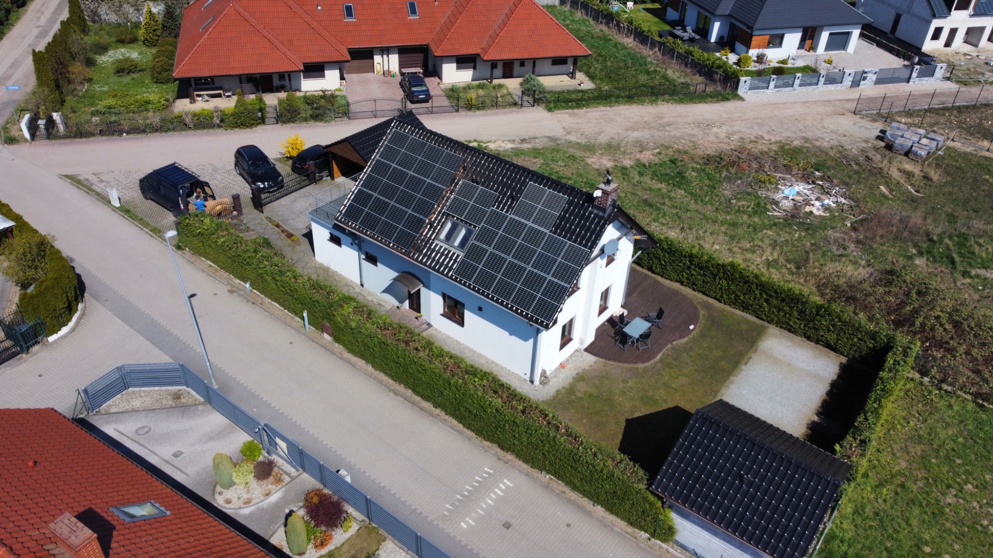 Widok z góry na biały dom z czarnymi panelami słonecznymi na dachu, otoczony zielonym żywopłotem, z zaparkowanymi samochodami i elementami ogrodu.