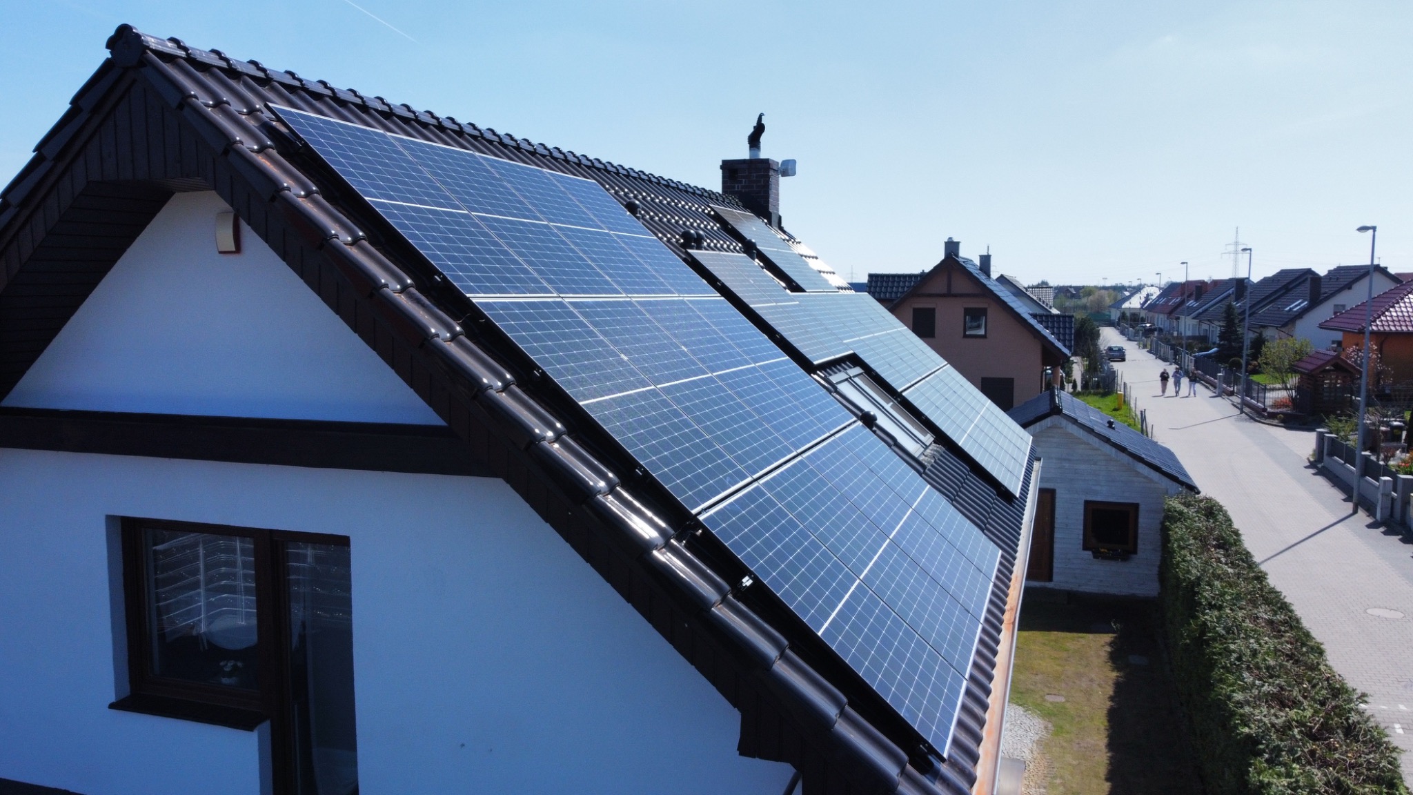 Panele fotowoltaiczne zamontowane na dachu domu jednorodzinnego z czarną dachówką, widok z góry na okoliczne domy i ulicę.