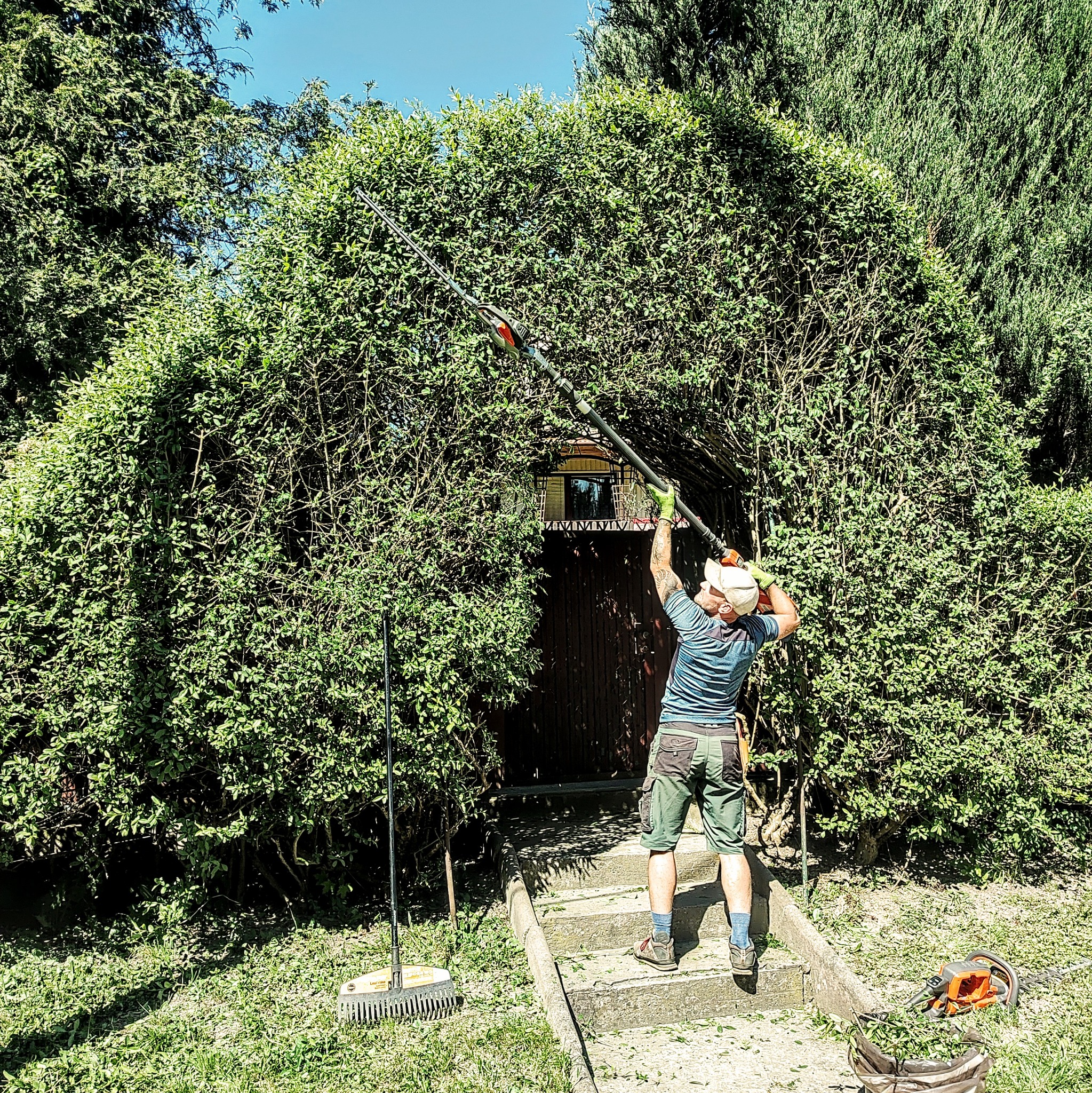 Mężczyzna w ogrodzie przycina wysoki żywopłot nad wejściem do domu za pomocą elektrycznych nożyc na wysięgniku; obok leżą grabie i nożyce do żywopłotu oraz worek z obciętymi gałązkami.