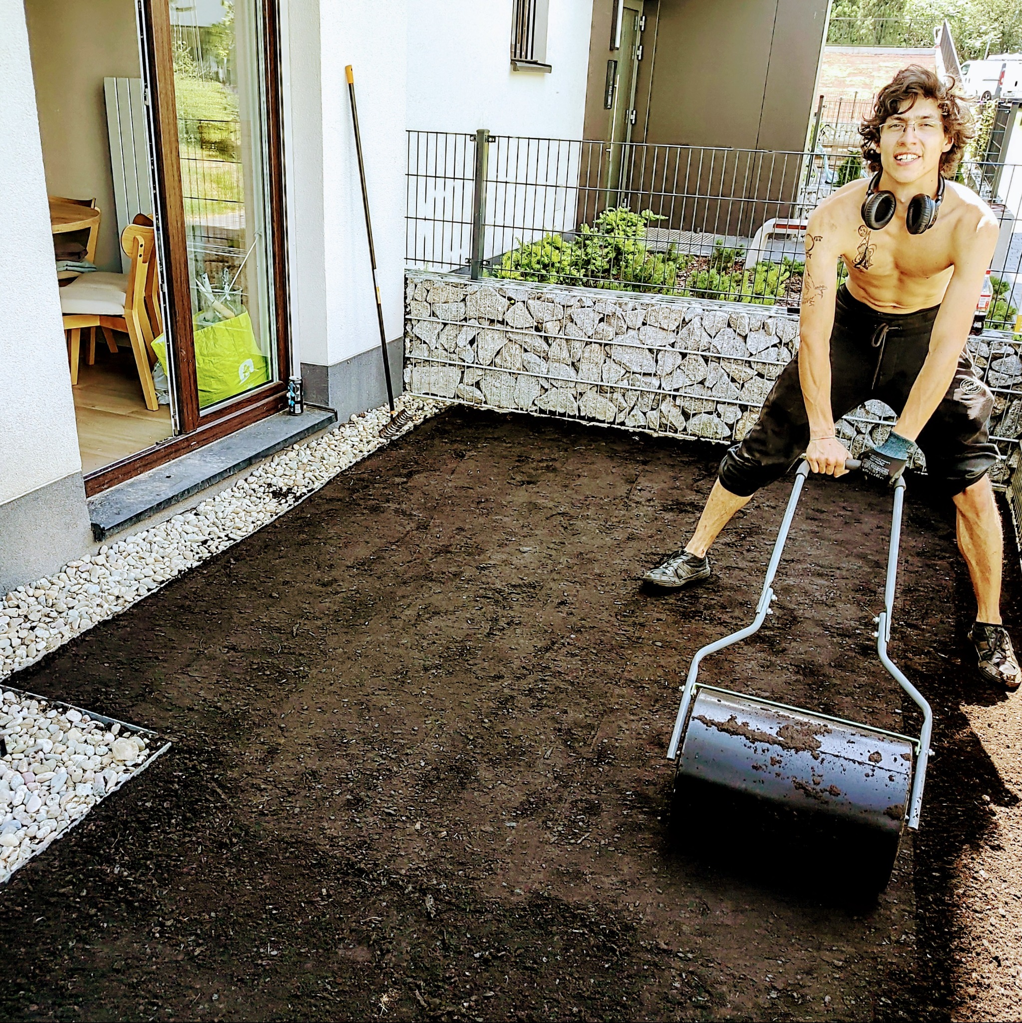 Mężczyzna bez koszulki ubija walcem ziemię w ogrodzie przydomowym, widoczny trawnik przygotowany pod zasiew lub rozłożenie trawy z rolki, w tle siatka gabionowa i budynek.