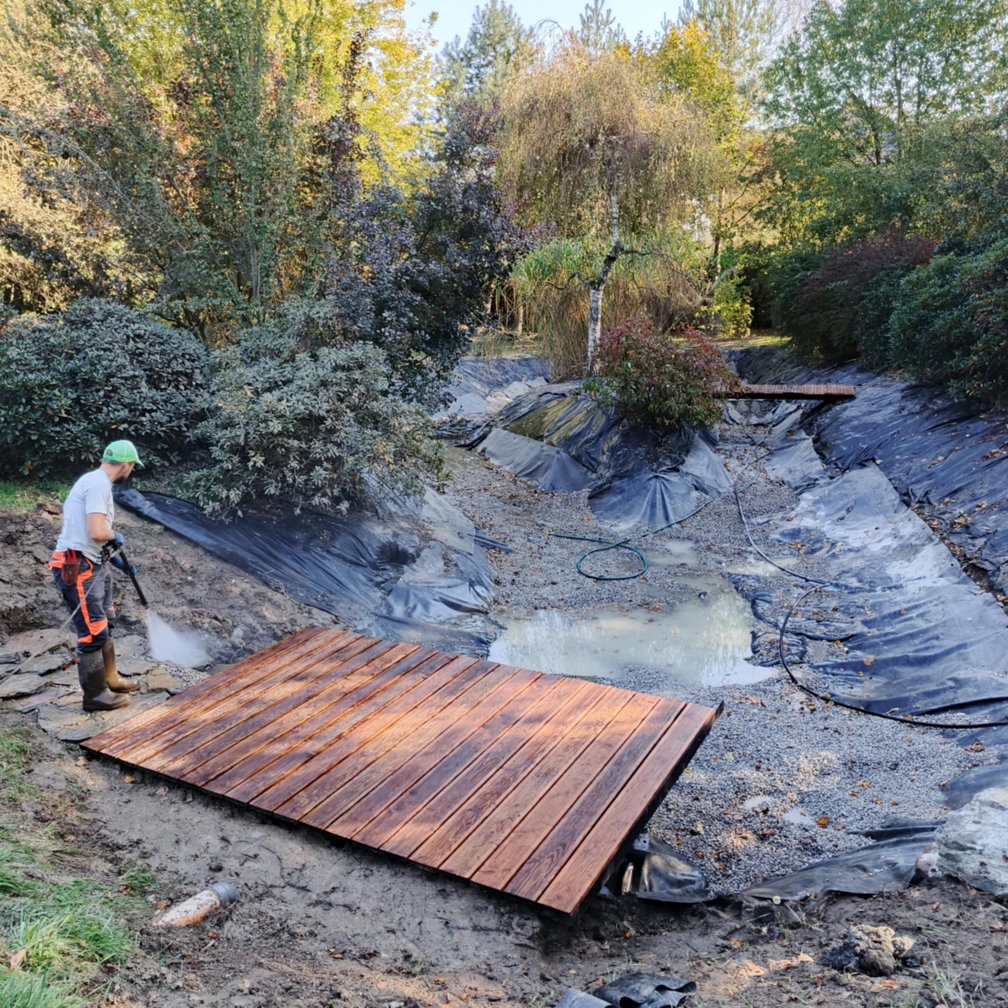 Pracownik w zielonej czapce czyści myjką ciśnieniową nowo zbudowany drewniany pomost nad wyłożonym czarną folią korytem stawu w ogrodzie.