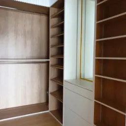 Wnętrze garderoby z otwartą częścią z półkami, zamkniętą przesuwnymi drzwiami z drewnopodobnego materiału oraz białą częścią z szufladami i lustrem w złotej ramie.