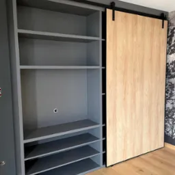 Szara szafa wnękowa z przesuwnymi drzwiami w kolorze jasnego drewna, zamontowana na czarnym systemie szynowym, na tle ściany z tapetą w motywy drzew.