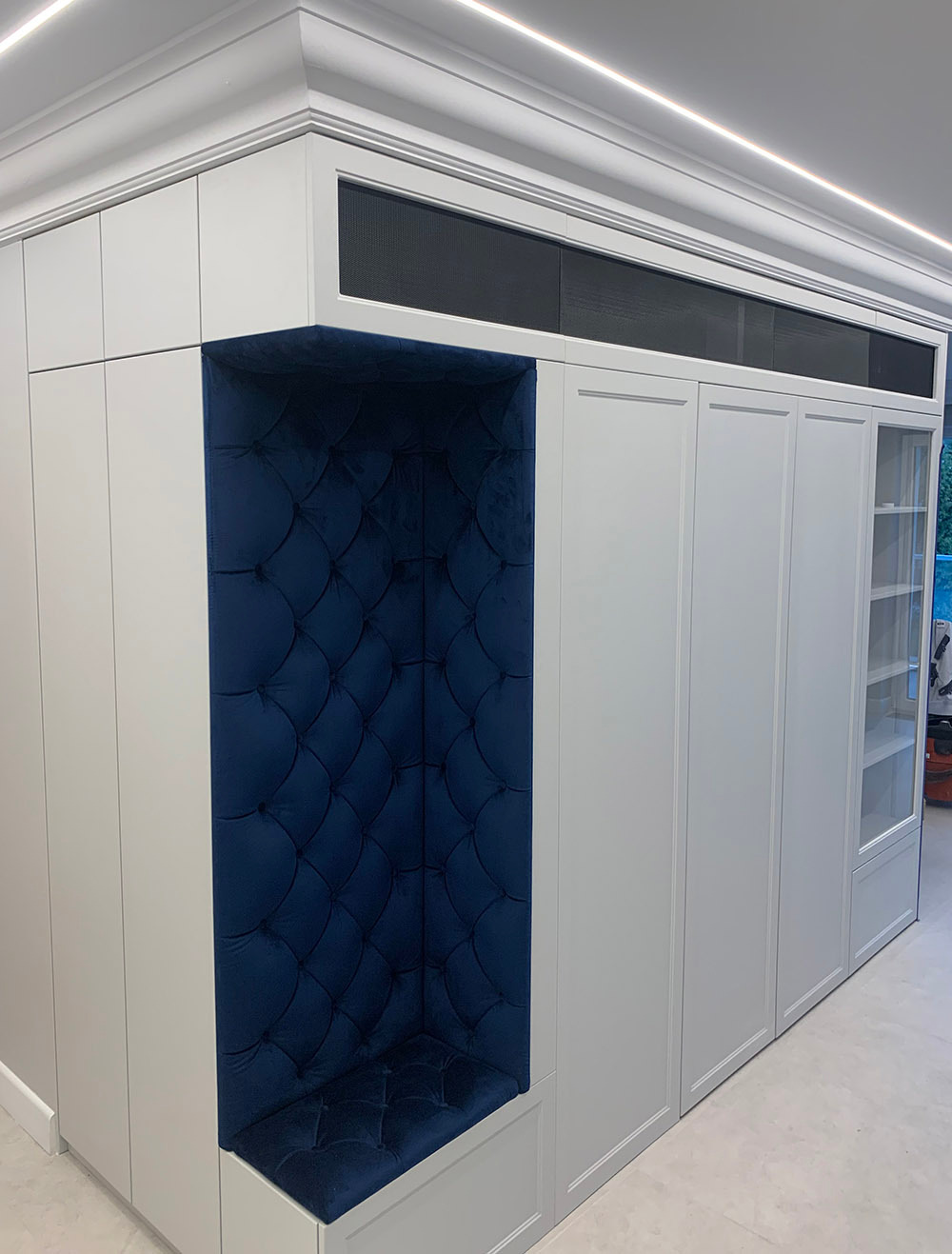 Biała zabudowa meblowa z wnęką wyściełaną granatowym, pikowanym welurem, tworząca siedzisko w przedpokoju, oświetlenie LED w suficie.