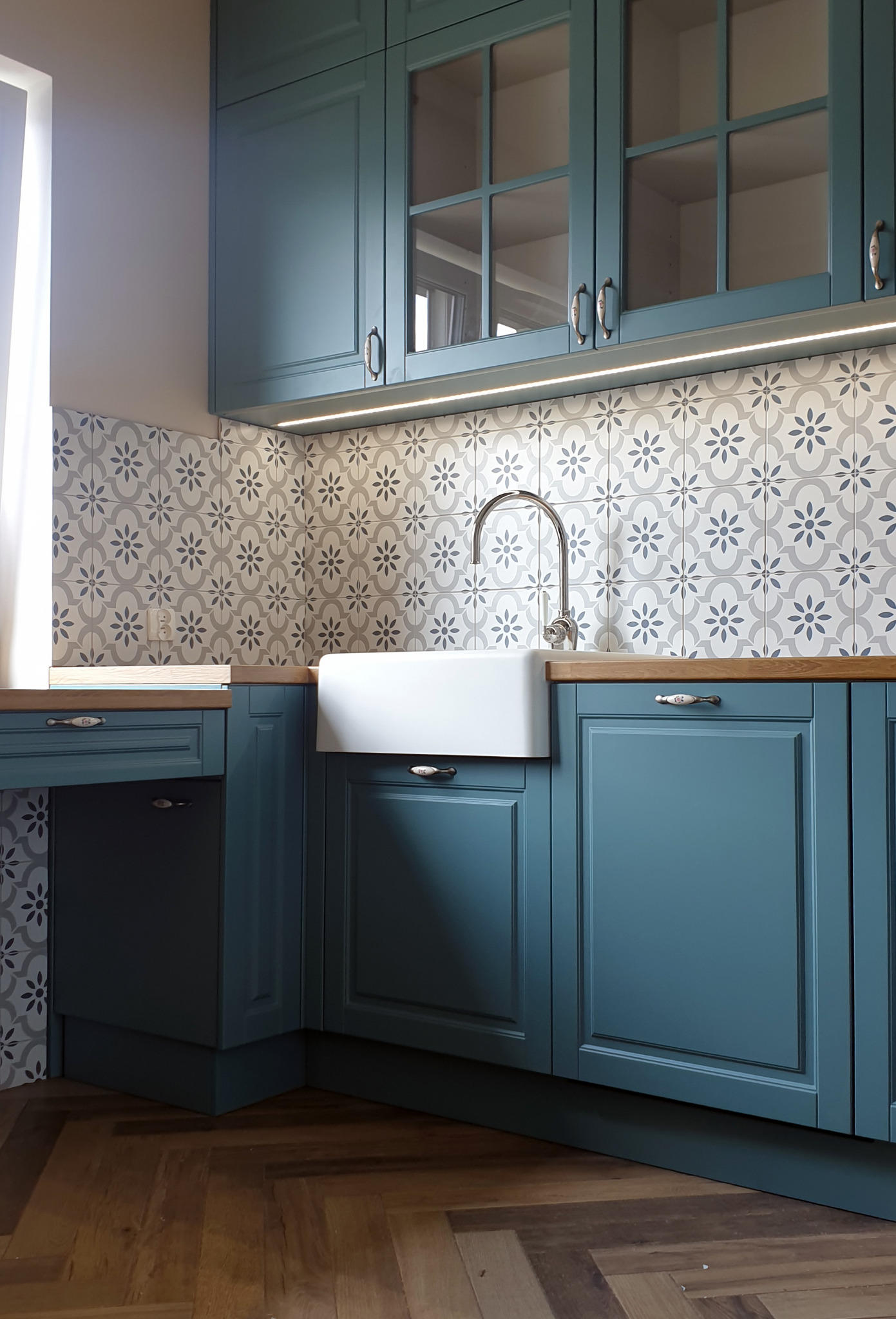 Turkusowa zabudowa kuchenna z frezowanymi frontami, ceramicznym zlewem farmerskim i baterią retro na tle ściany z dekoracyjnymi płytkami w kwiatowy wzór, oświetlona listwą LED pod szafkami wiszącymi.