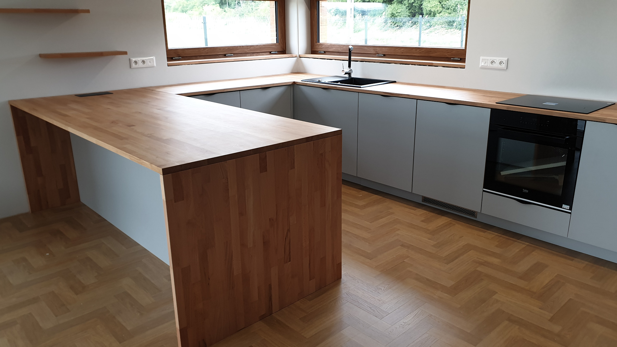 Nowoczesna kuchnia z szarymi szafkami, drewnianym blatem i wyspą, uzupełniona o czarną armaturę i sprzęt AGD, na parkiecie w jodełkę.