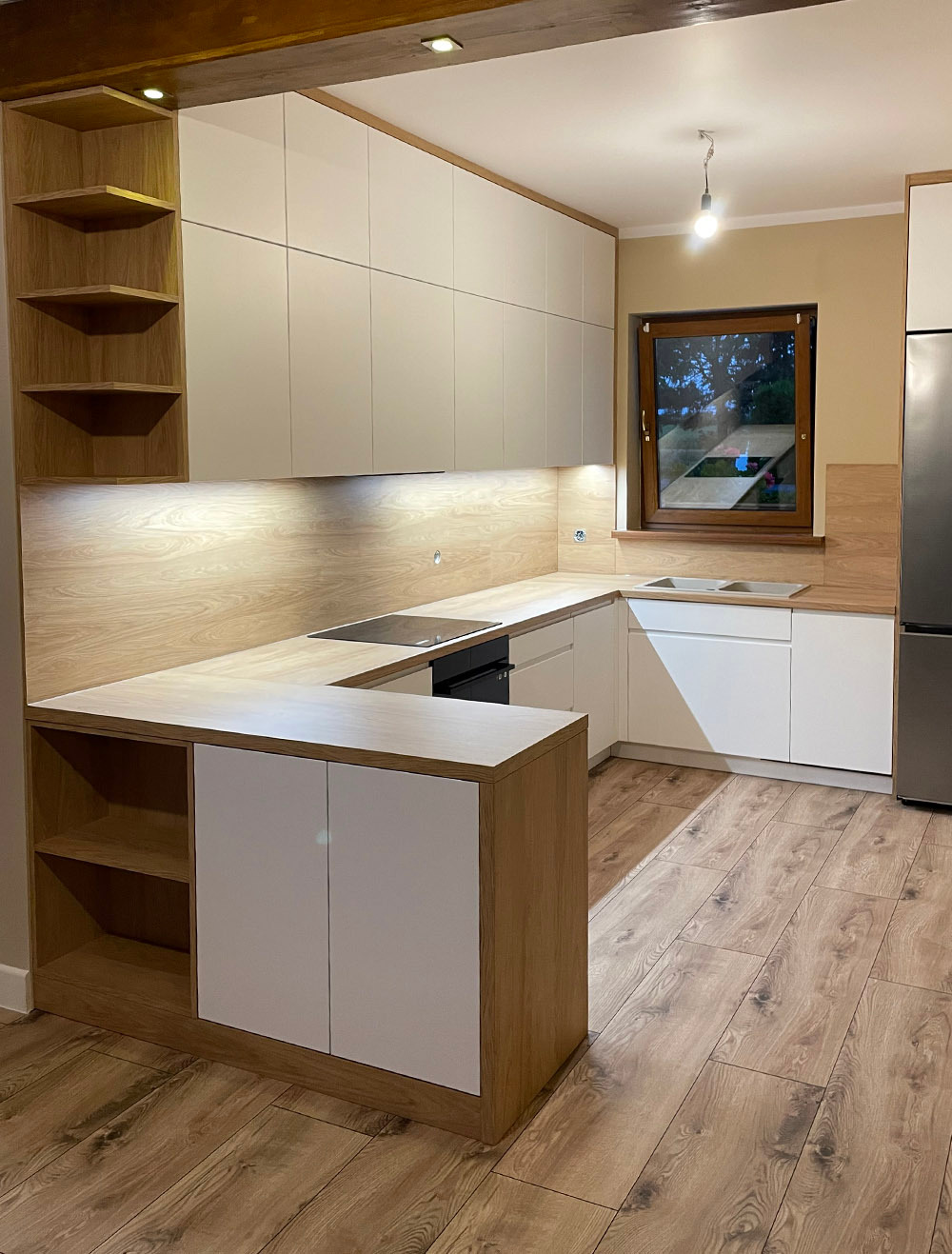 Nowoczesna kuchnia w kształcie litery U z jasnymi frontami szafek, drewnianymi blatami i otwartymi półkami z boku, oświetlona listwą LED pod szafkami wiszącymi i pojedynczą lampą wiszącą.