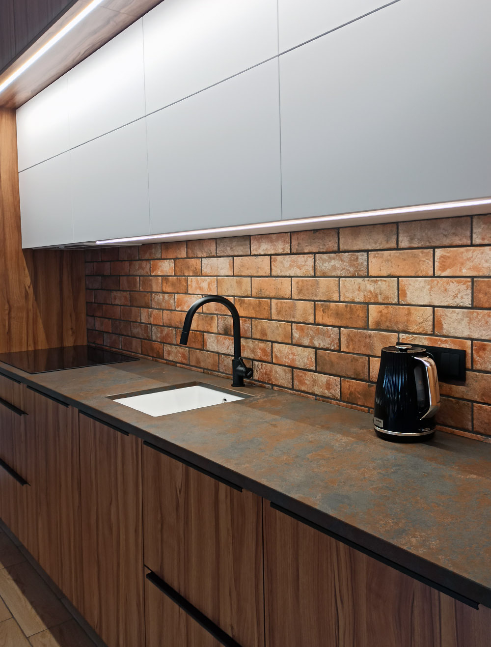Nowoczesna kuchnia z drewnianymi szafkami, blatem w odcieniach rdzy i ceglaną ścianą, oświetlona listwą LED pod górnymi szafkami. Na blacie czarny czajnik elektryczny i minimalistyczna, biała...