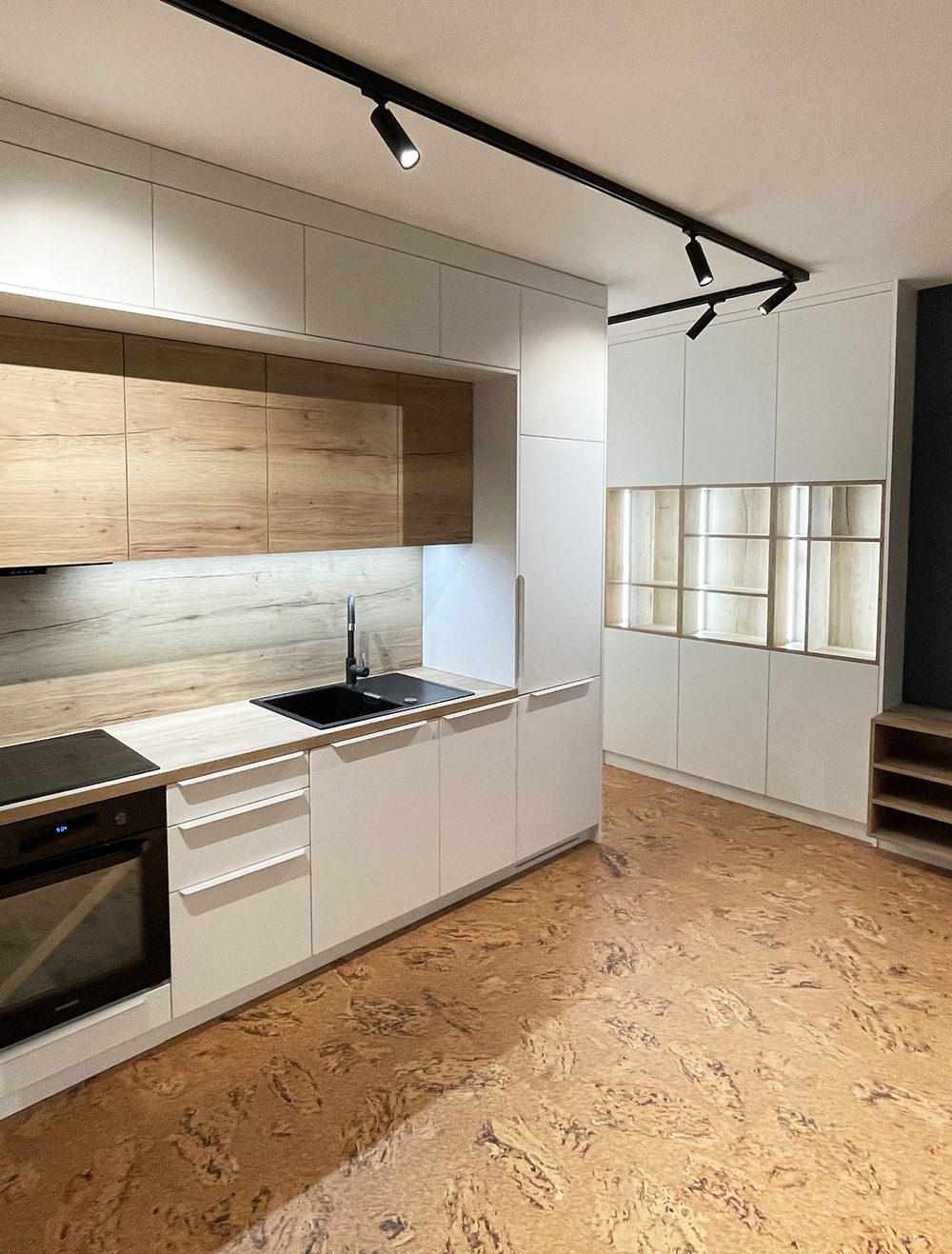 Nowoczesna kuchnia z białymi szafkami i drewnianymi akcentami, czarnym zlewem i płytą indukcyjną, oświetleniem punktowym na szynie i podłogą z korka.