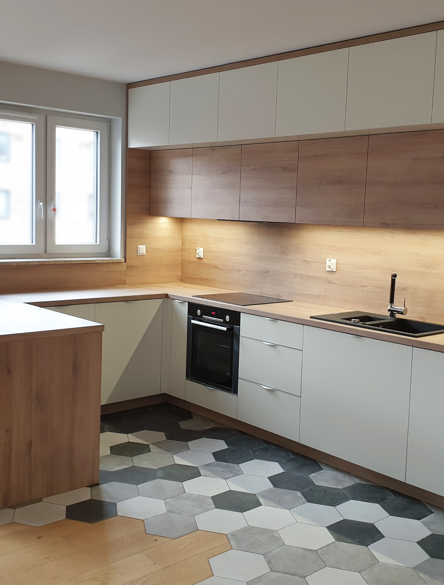 Nowoczesna kuchnia z białymi i drewnopodobnymi frontami, sześciokątne płytki podłogowe w odcieniach szarości, czarny zlewozmywak i płyta indukcyjna, oświetlenie podszafkowe.