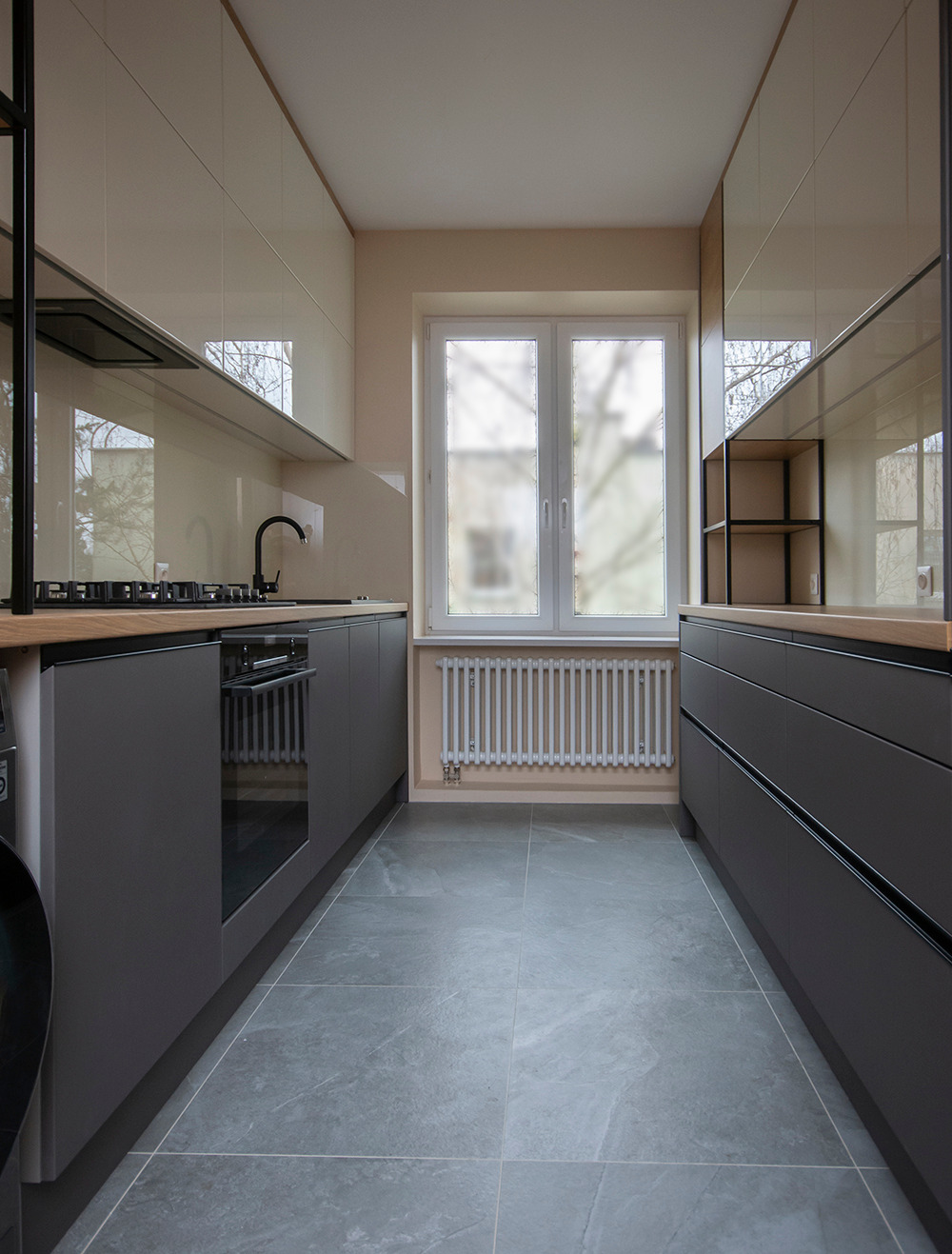 Wąska, nowoczesna kuchnia z szarymi szafkami dolnymi i kremowymi górnymi, drewnianym blatem, czarną armaturą i szarą podłogą z płytek, oświetlona naturalnym światłem z okna.