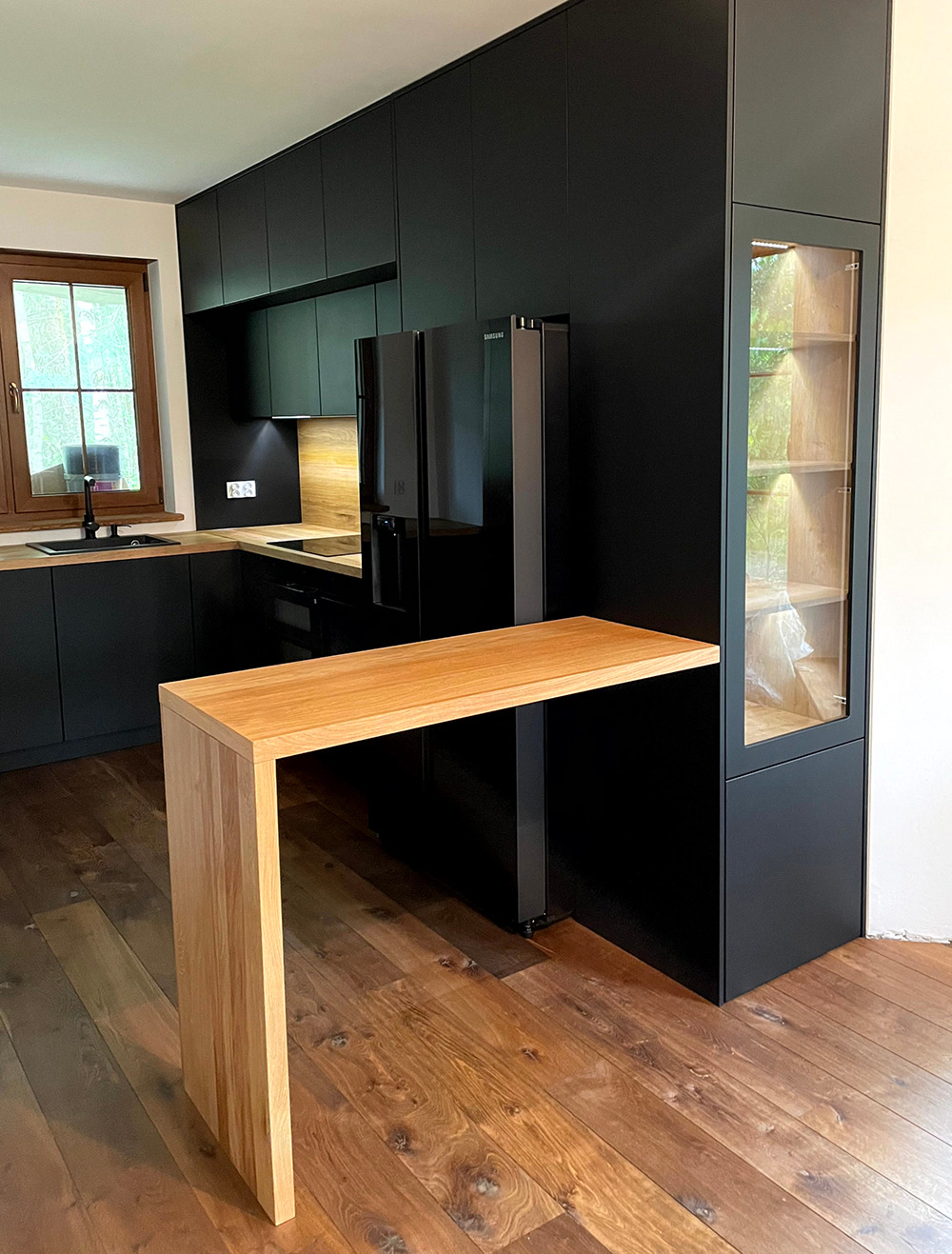 Nowoczesna kuchnia z czarnymi szafkami i drewnianym blatem, uzupełniona o wysoki, drewniany stół barowy z litego drewna, podkreślona ciepłym oświetleniem i drewnianą podłogą.