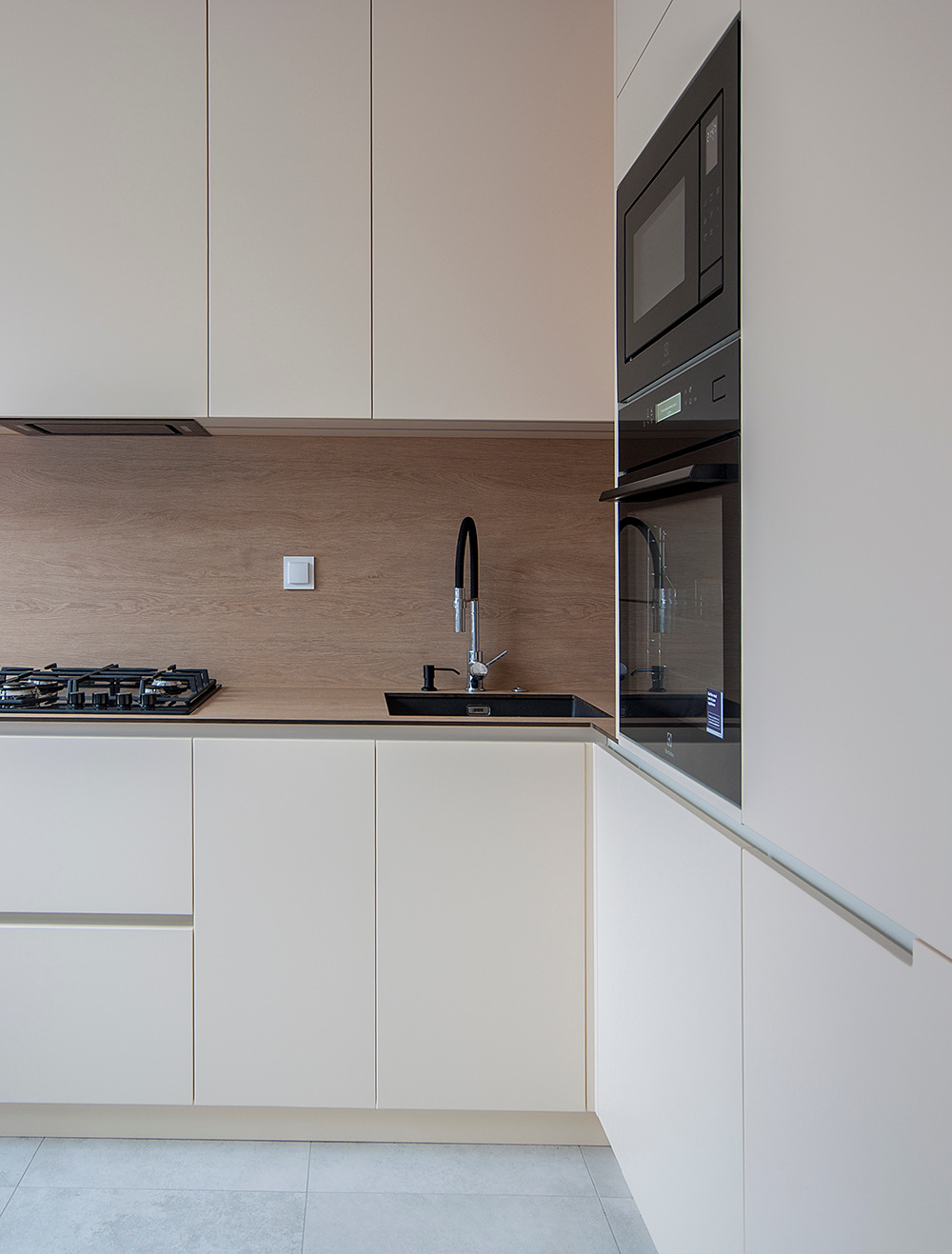 Jasna kuchnia z gładkimi frontami, czarną armaturą i zabudowanym piekarnikiem z mikrofalówką, widok na płytę gazową i drewnopodobny panel ścienny.