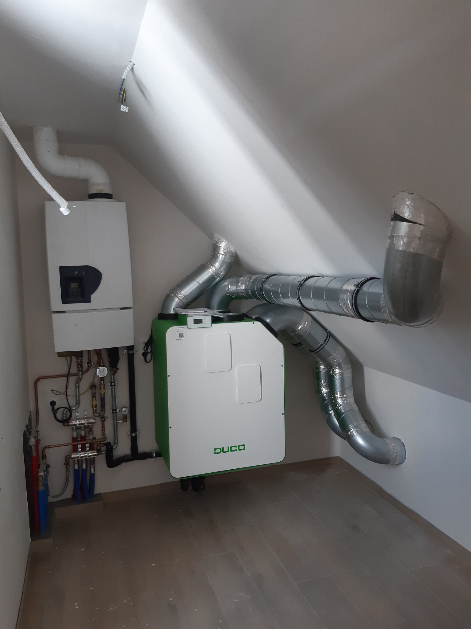 Biała centrala wentylacyjna z zielonymi akcentami marki Duco, podłączona do systemu rur wentylacyjnych w pomieszczeniu na poddaszu, obok kotła grzewczego i instalacji hydraulicznej.