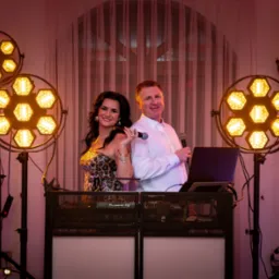 Duet muzyczny: kobieta w cekinowej sukience i mężczyzna w koszuli za konsoletą DJ-ską, otoczeni światłami na statywach. Uśmiechnięci, gotowi do występu.