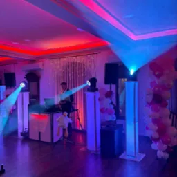 Sala z DJ-em podczas imprezy, oświetlona czerwonymi i niebieskimi światłami. Dekoracje z balonów i pluszowa maskotka dodają uroku.