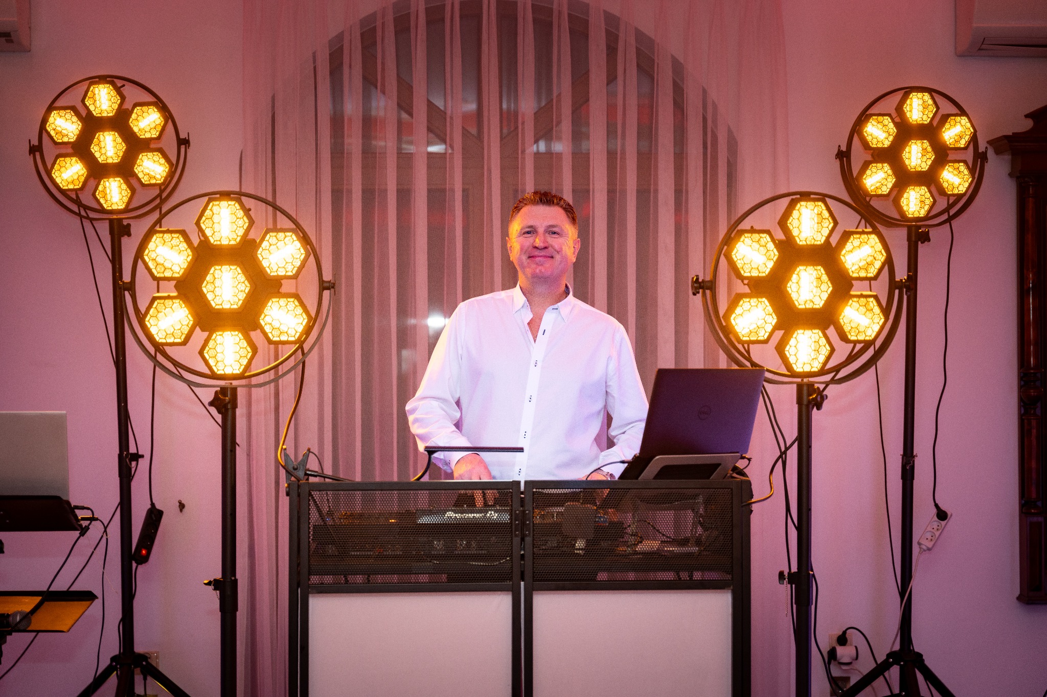 DJ w białej koszuli za konsoletą Pioneer DJ, otoczony przez dwa reflektory z ciepłym światłem, w tle okno z zasłonami, tworząc klimat imprezy.