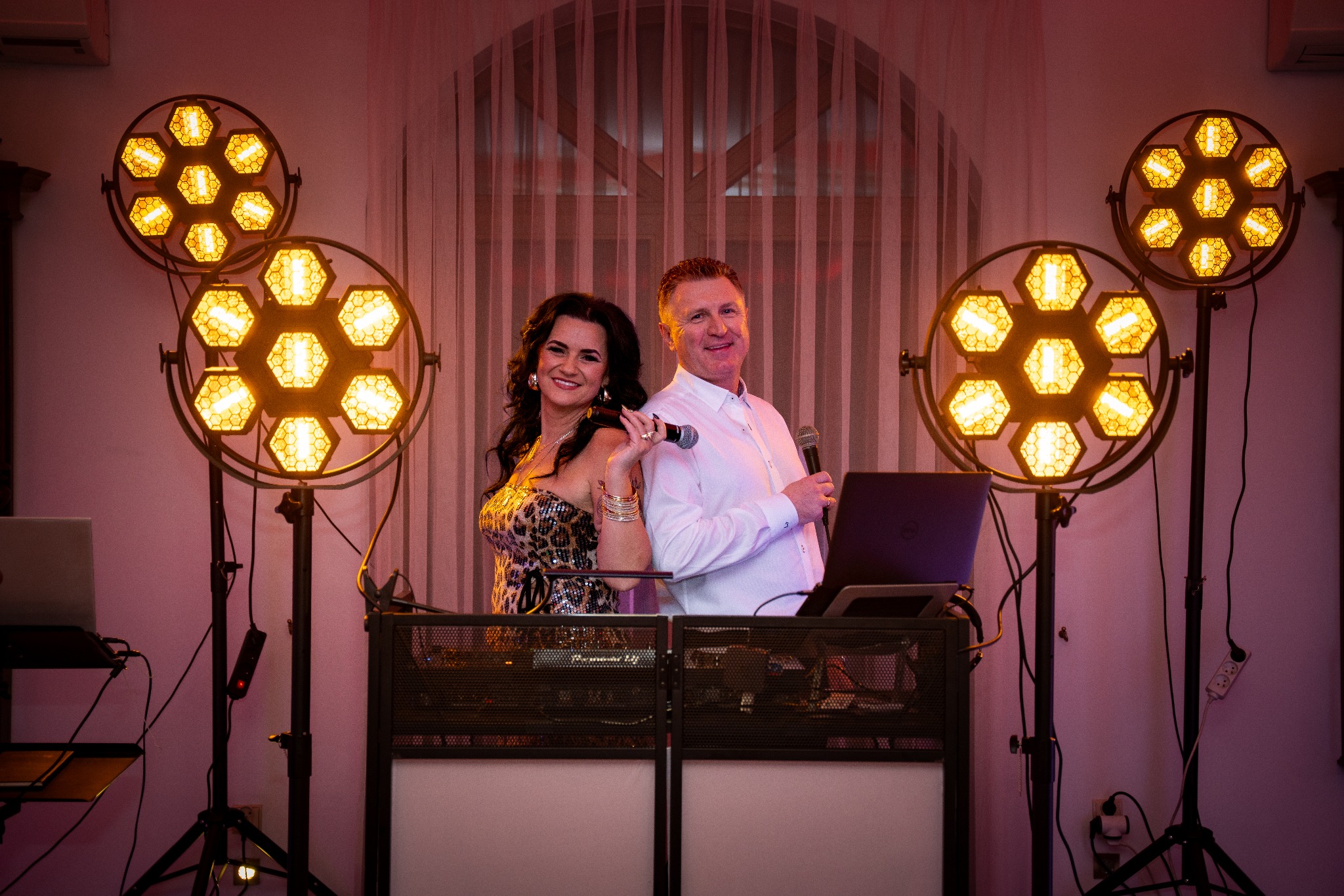 Duet muzyczny: kobieta w cekinowej sukience i mężczyzna w koszuli za konsoletą DJ-ską, otoczeni światłami na statywach. Uśmiechnięci, gotowi do występu.