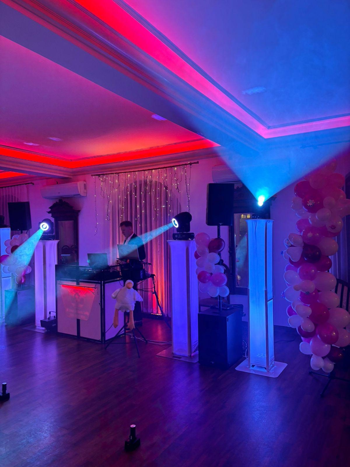 Sala z DJ-em podczas imprezy, oświetlona czerwonymi i niebieskimi światłami. Dekoracje z balonów i pluszowa maskotka dodają uroku.
