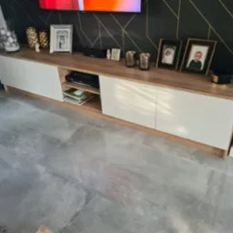 Nowoczesna szafka RTV z białymi frontami i blatem w kolorze drewna, na kt&oacute;rej stoją ramki ze zdjęciami, wazony i świeczki, na tle ściany z geometrycznym wzorem i szarych płytek podłogowych.