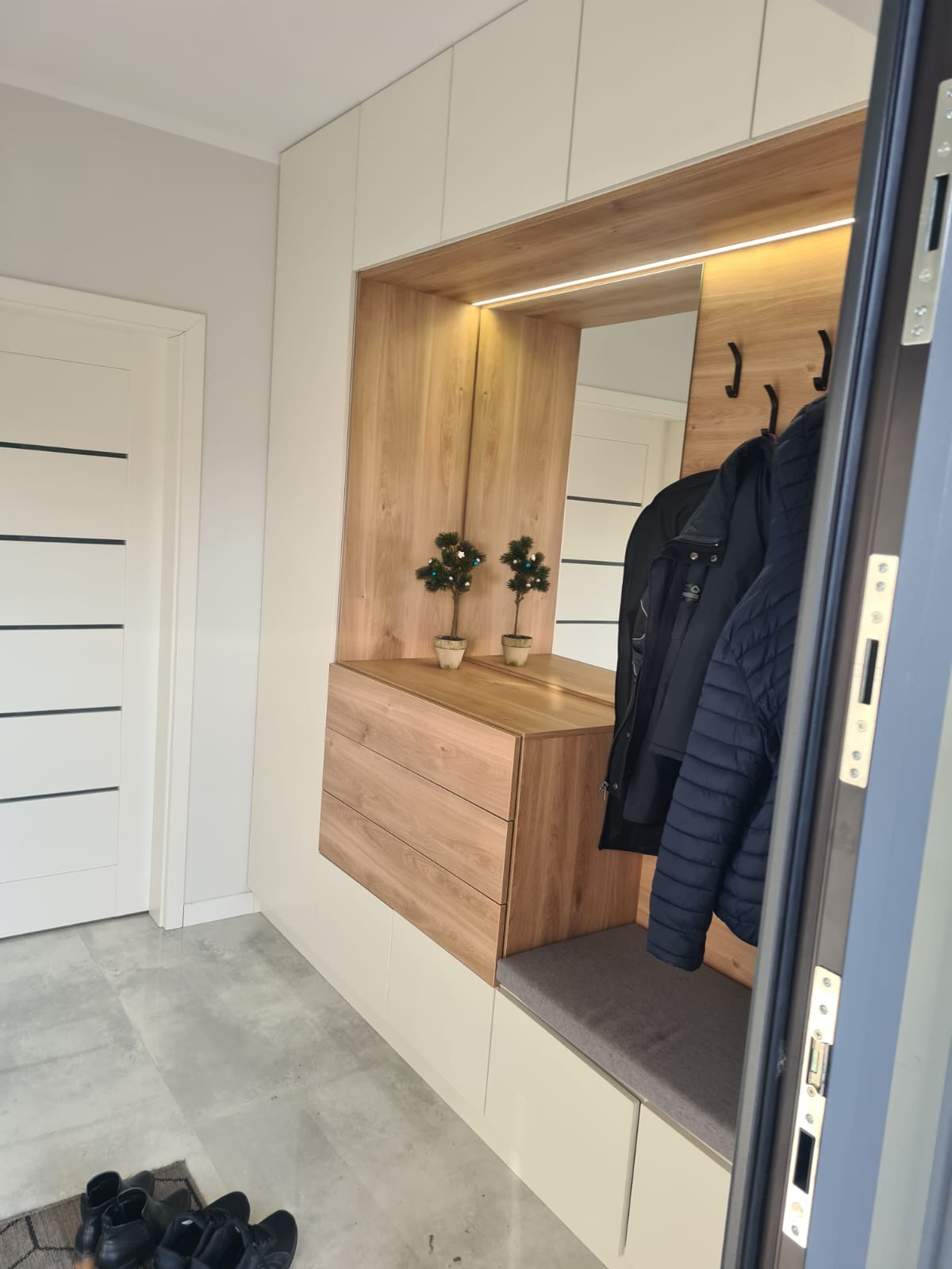 Nowoczesna szafa wnękowa z drewnianymi frontami i oświetleniem LED, z siedziskiem i wieszakami na ubrania, dekorowana dwoma małymi drzewkami świątecznymi.