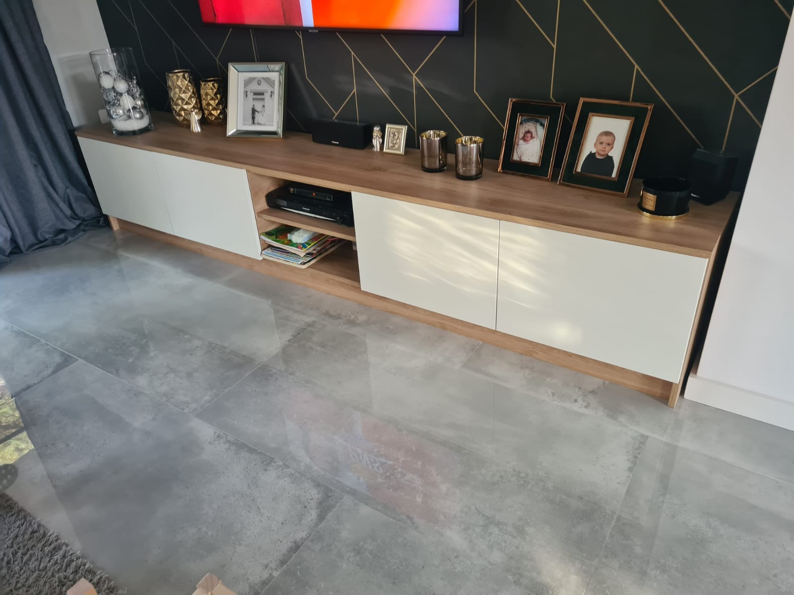 Nowoczesna szafka RTV z białymi frontami i blatem w kolorze drewna, na której stoją ramki ze zdjęciami, wazony i świeczki, na tle ściany z geometrycznym wzorem i szarych płytek podłogowych.