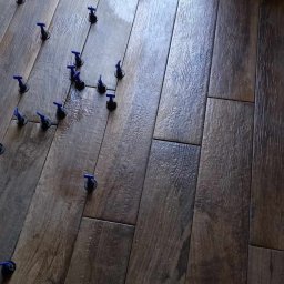 EMa - Miłosz Zadka - Ułożone, brązowe panele podłogowe imitujące drewno, z niebieskimi elementami poziomującymi, tworzą wzór w jasnym wnętrzu. Widok z góry, ukośny kadr.