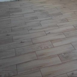 EMa - Miłosz Zadka - Ułożone, podłużne płytki podłogowe imitujące drewno, z widocznymi fugami. Płytki w odcieniach beżu i brązu, ułożone równolegle do ściany. Widoczny fragment ściany.