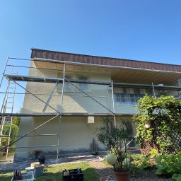 EMa - Miłosz Zadka - Elewacja domu w trakcie malowania, rusztowanie, zabezpieczone okna folią, widoczne narzędzia malarskie, słoneczny dzień.