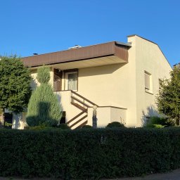 EMa - Miłosz Zadka - Jasny, dwukondygnacyjny dom z brązowym zadaszeniem nad balkonem i drewnianymi schodami wejściowymi, otoczony zielenią w słoneczny dzień. Widok z poziomu ulicy.