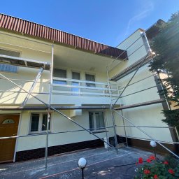 EMa - Miłosz Zadka - Dom jednorodzinny w trakcie odświeżania elewacji, otoczony rusztowaniem. Widoczne świeżo pomalowane ściany w jasnym odcieniu i elementy zabezpieczające teren prac.