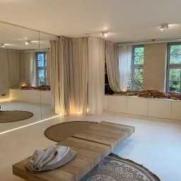 EMa - Miłosz Zadka - Jasne, minimalistyczne wnętrze w Kraszewicach z drewnianym stolikiem, okrągłymi dywanami i zasłonami. Ściany w odcieniach beżu, duże okna z widokiem na zieleń.