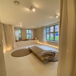 EMa - Miłosz Zadka - Jasne, minimalistyczne wnętrze z drewnianym podestem, okrągłymi dywanami i zwiewnymi zasłonami. Subtelne oświetlenie LED podkreśla nowoczesny charakter pomieszczenia.