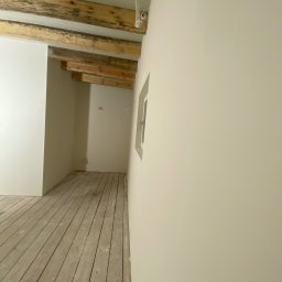 EMa - Miłosz Zadka - Jasne wnętrze po odświeżeniu: pomalowane ściany, drewniana podłoga i belki stropowe. Widoczne okno i instalacja elektryczna.