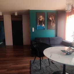 EMa - Miłosz Zadka - Salon po metamorfozie: drewniana podłoga, turkusowa ściana z obrazami, szara sofa i okrągły stolik z marmurowym blatem. Nowoczesne lampy i dekoracje.
