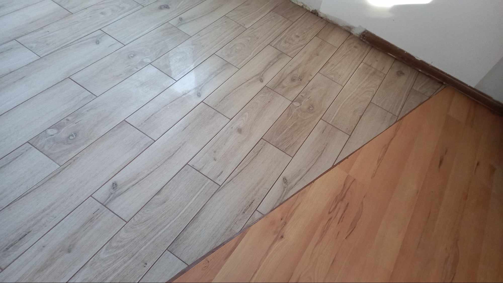 Fragment podłogi z ułożonymi panelami w odcieniach szarości, kontrastujący z fragmentem starej, brązowej podłogi. Widoczny narożnik ściany.