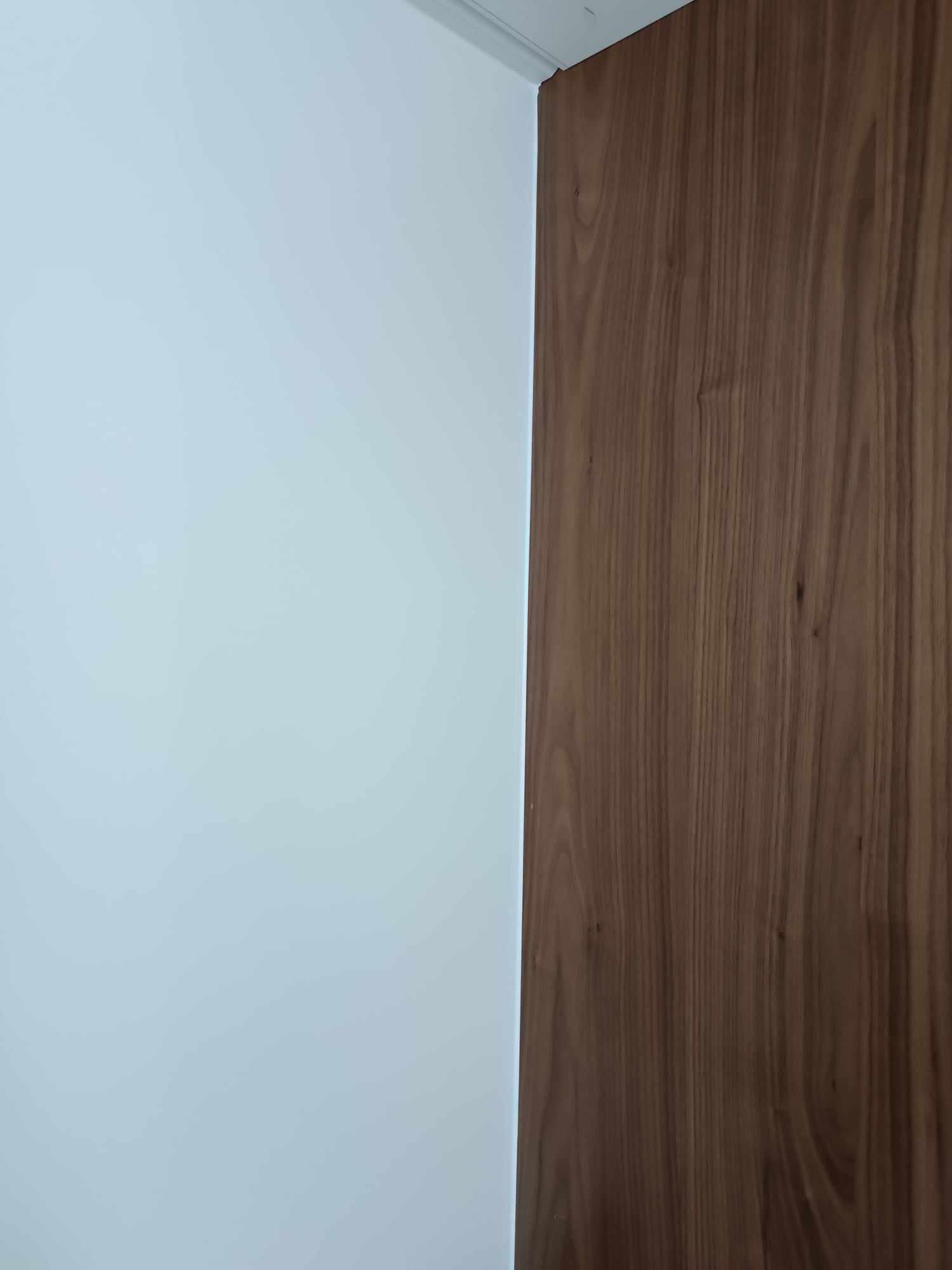 Biała ściana sąsiaduje z drewnianą okładziną ścienną w ciepłym odcieniu brązu. Minimalistyczne wykończenie wnętrza, widoczny fragment listwy przypodłogowej.