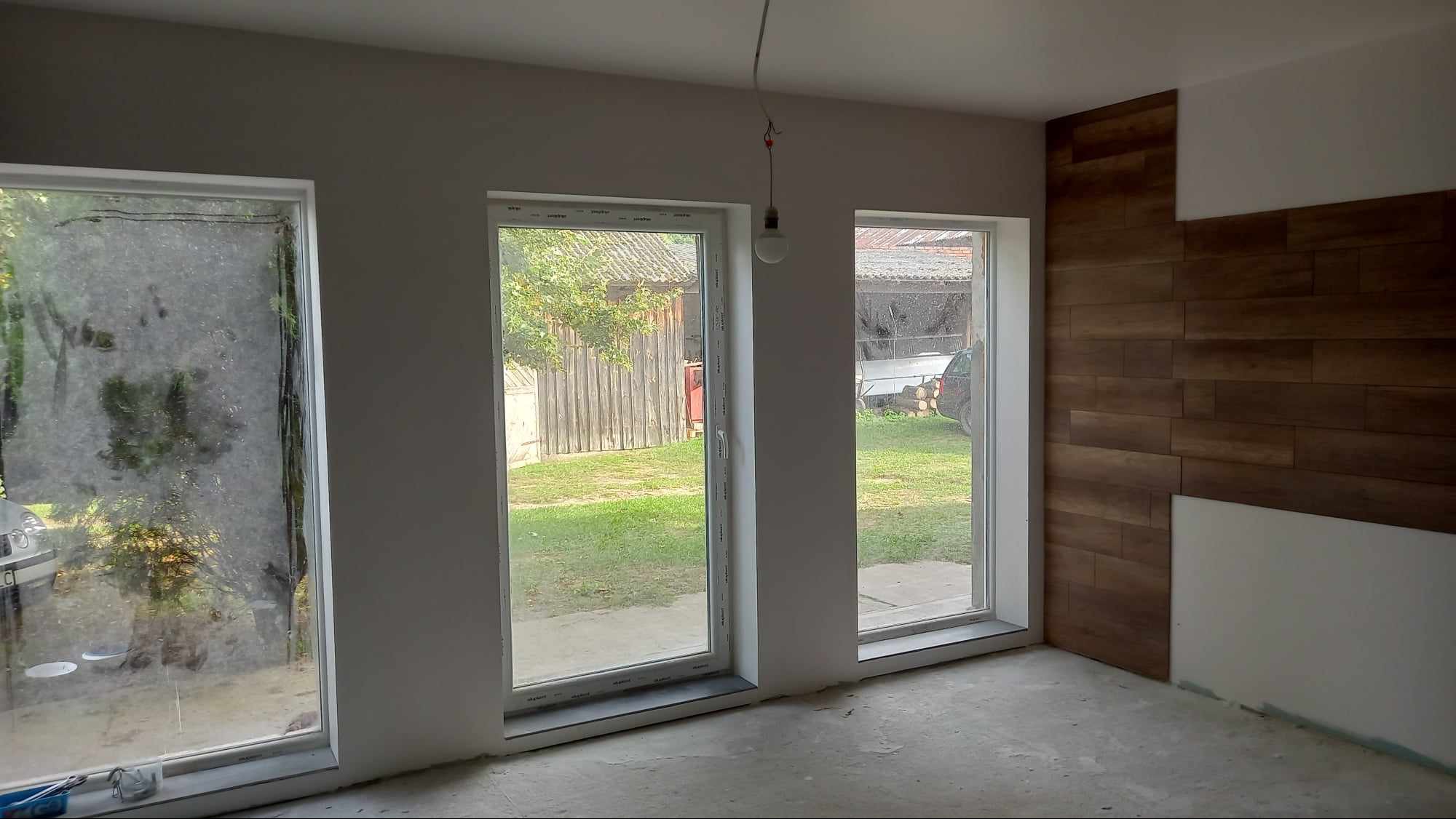 Wnętrze pokoju w trakcie prac wykończeniowych: częściowo ułożona okładzina ścienna imitująca drewno, surowe ściany i okna z widokiem na zewnątrz.