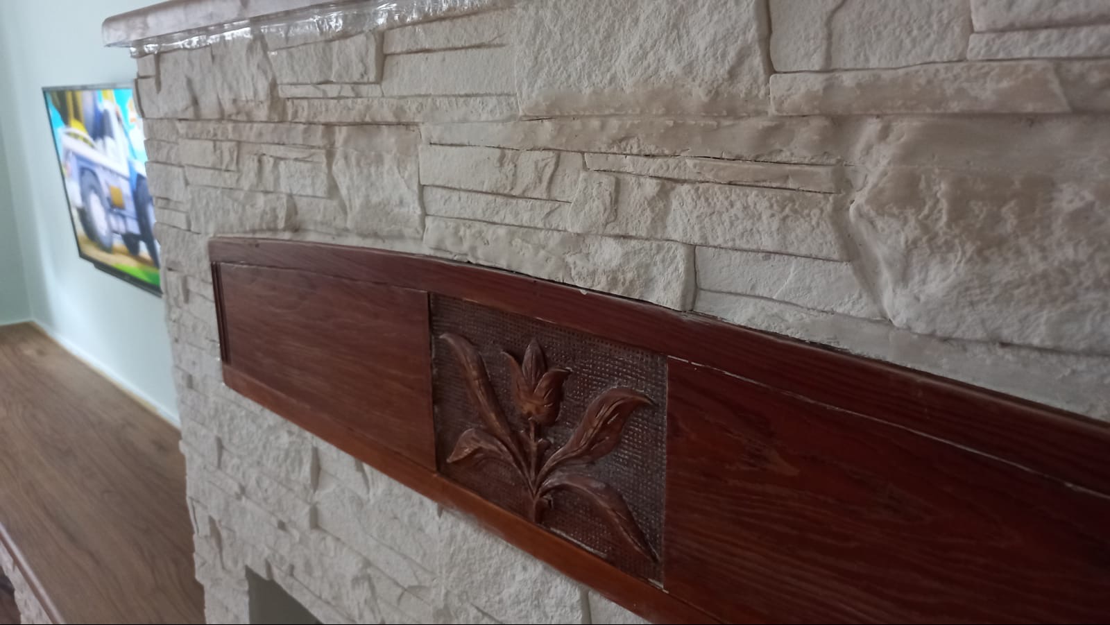 Dekoracyjna ściana z białego kamienia z drewnianym elementem ozdobnym z motywem roślinnym. W tle widoczny telewizor i fragment podłogi.