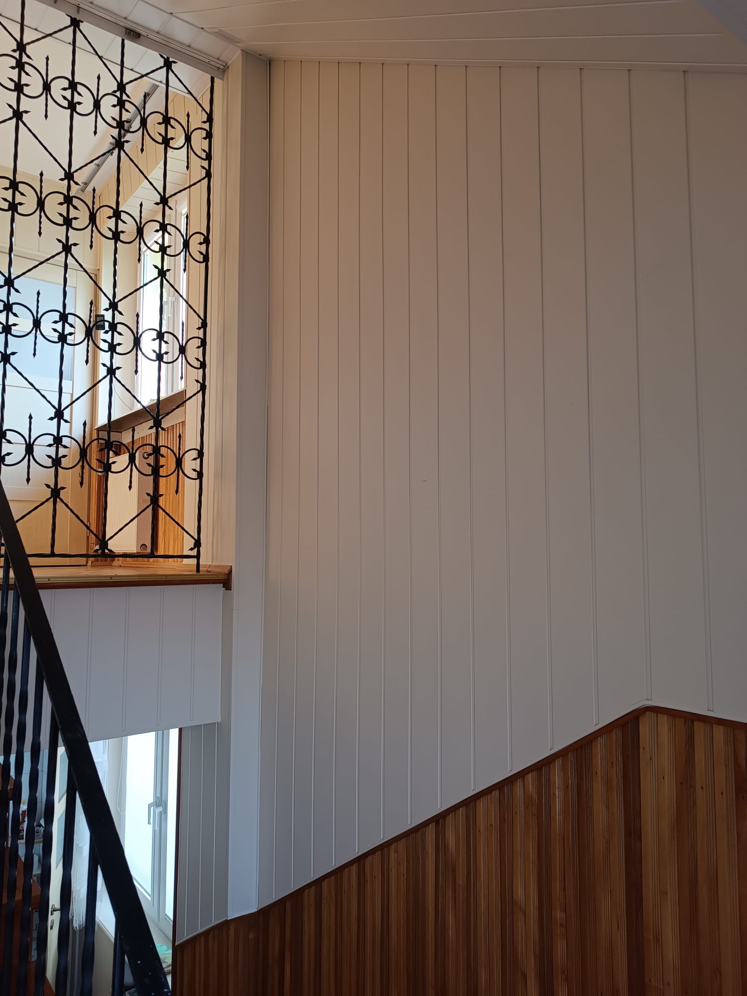 Białe panele ścienne z drewnianą boazerią u dołu, widoczne przez ozdobną, metalową balustradę schodów. Jasne wnętrze, pionowe linie paneli.