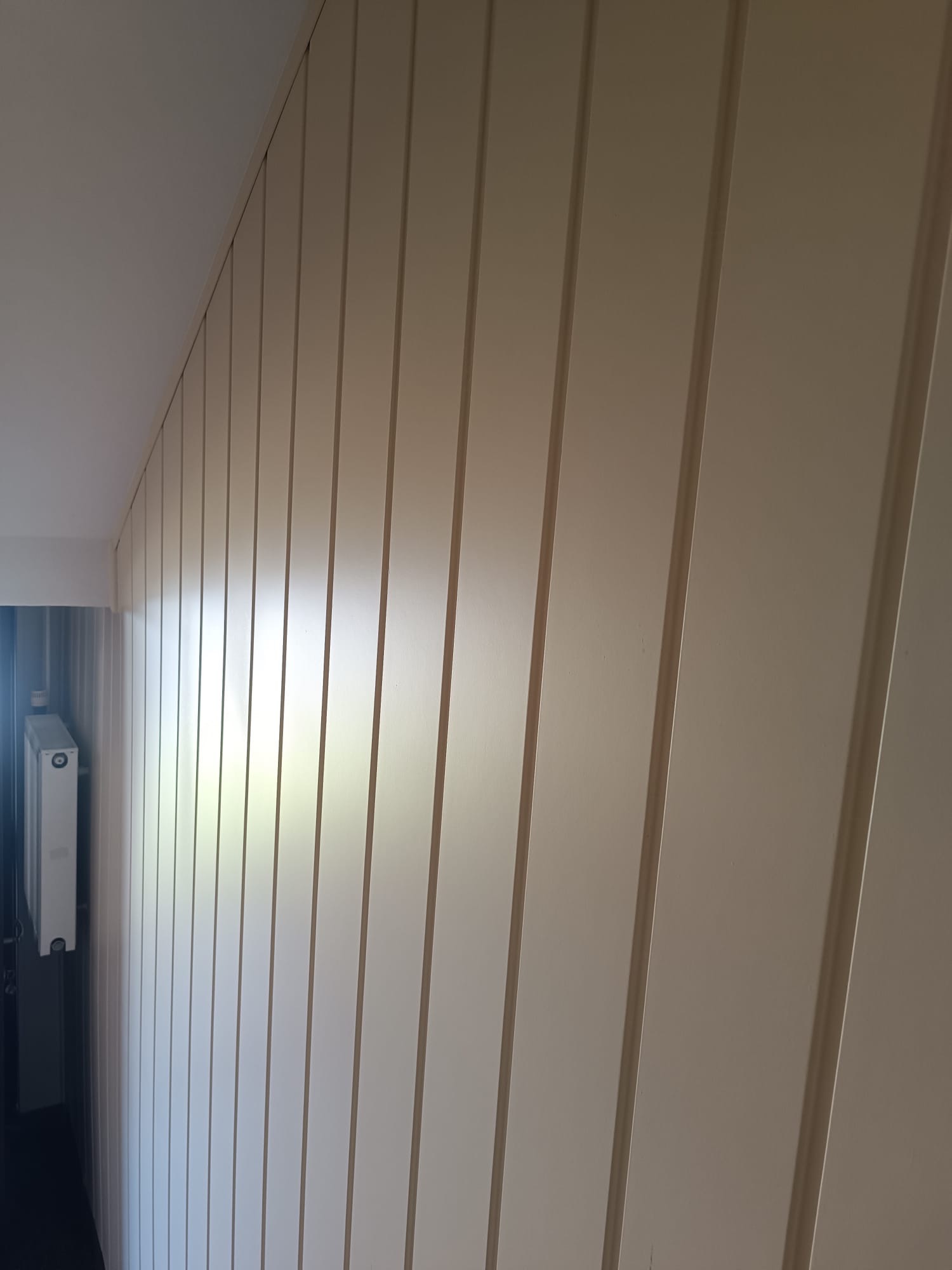 Biała, pionowa boazeria na ścianie, z widocznym fragmentem grzejnika w tle. Ujęcie z dołu, podkreślające strukturę i wykończenie ściany. Neutralna kolorystyka.