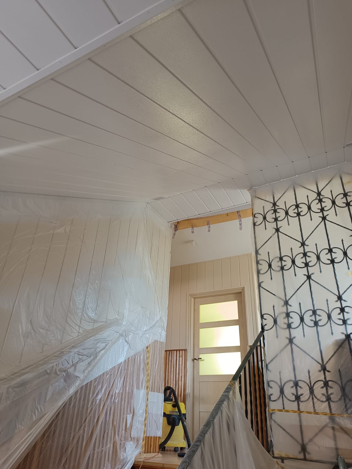 Biały, świeżo pomalowany sufit z boazerią, zabezpieczone folią ściany i schody. Widoczny odkurzacz przemysłowy. Ujęcie z dołu, w tle drzwi.