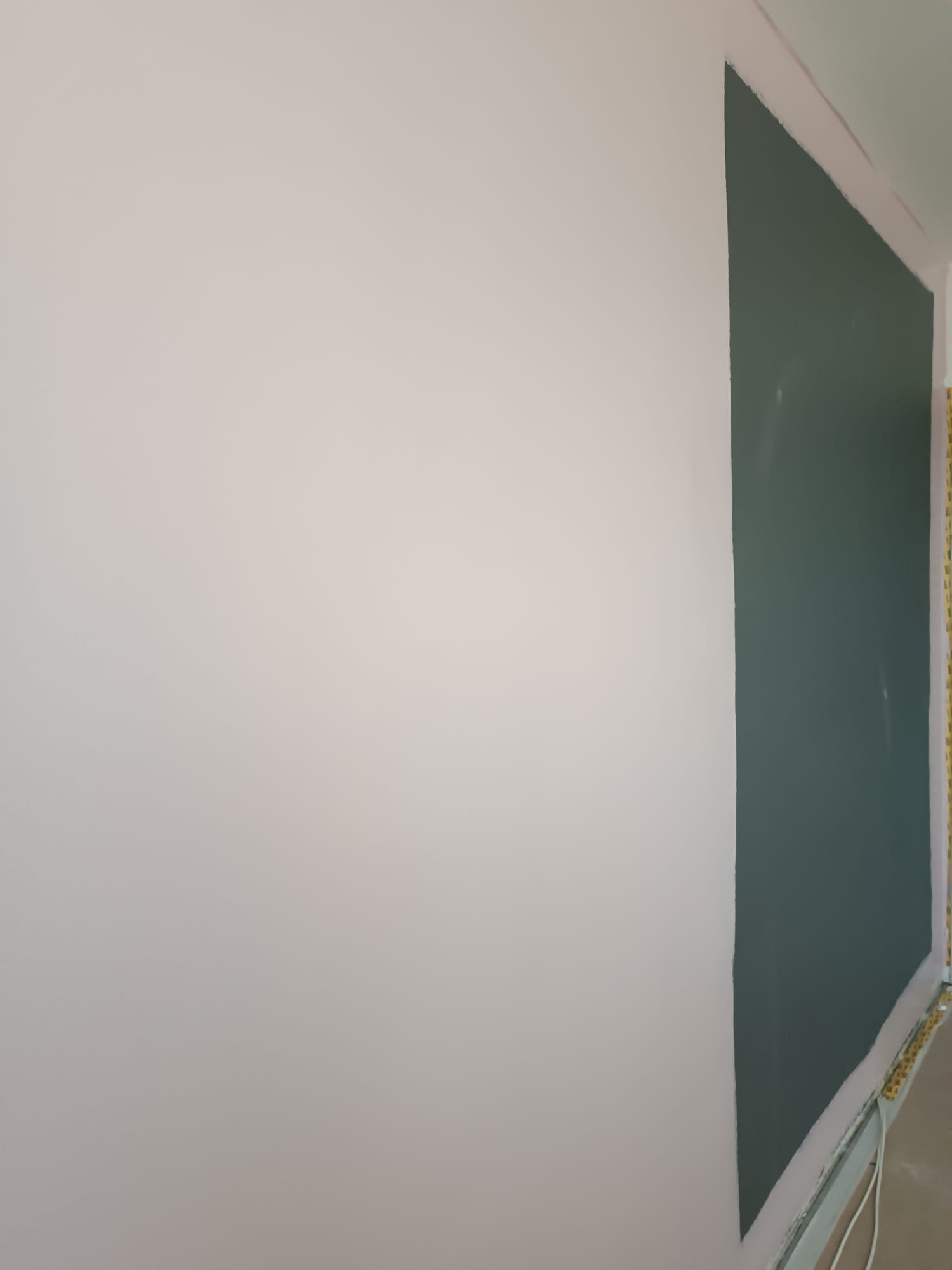 Ściana po częściowym pomalowaniu na dwa kolory: jasny beż i ciemny zielony. Widoczne taśmy malarskie przy suficie i podłodze. Ujęcie z boku, perspektywa z dołu.